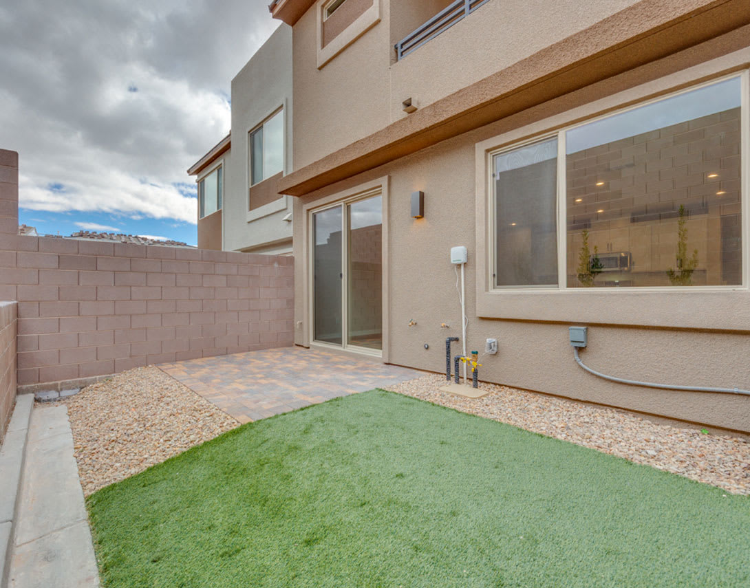 A modern townhouse with a small patio area featuring a grassy patch and a paved walkway, set against a cloudy sky backdrop.