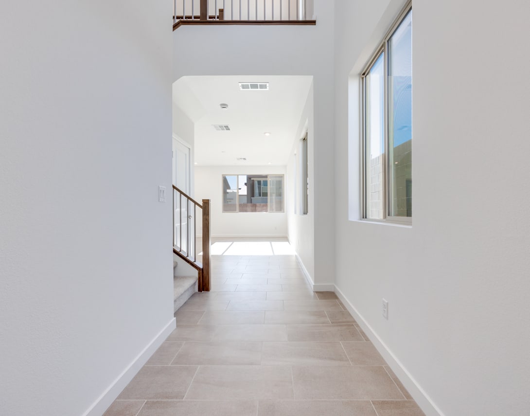 A bright, spacious hallway with white walls, a tiled floor, and a window at the end, providing natural light and a view of the outdoors.