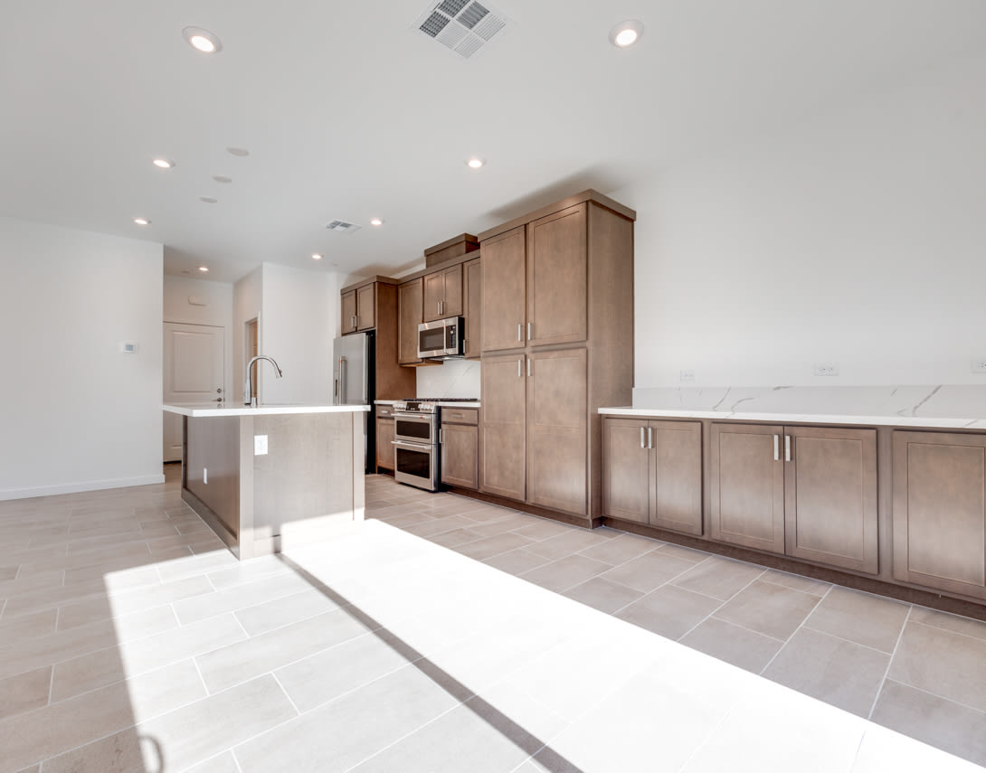 A modern, open-concept kitchen with wooden cabinets, stainless steel appliances, and a tiled floor, bathed in natural light from the windows.