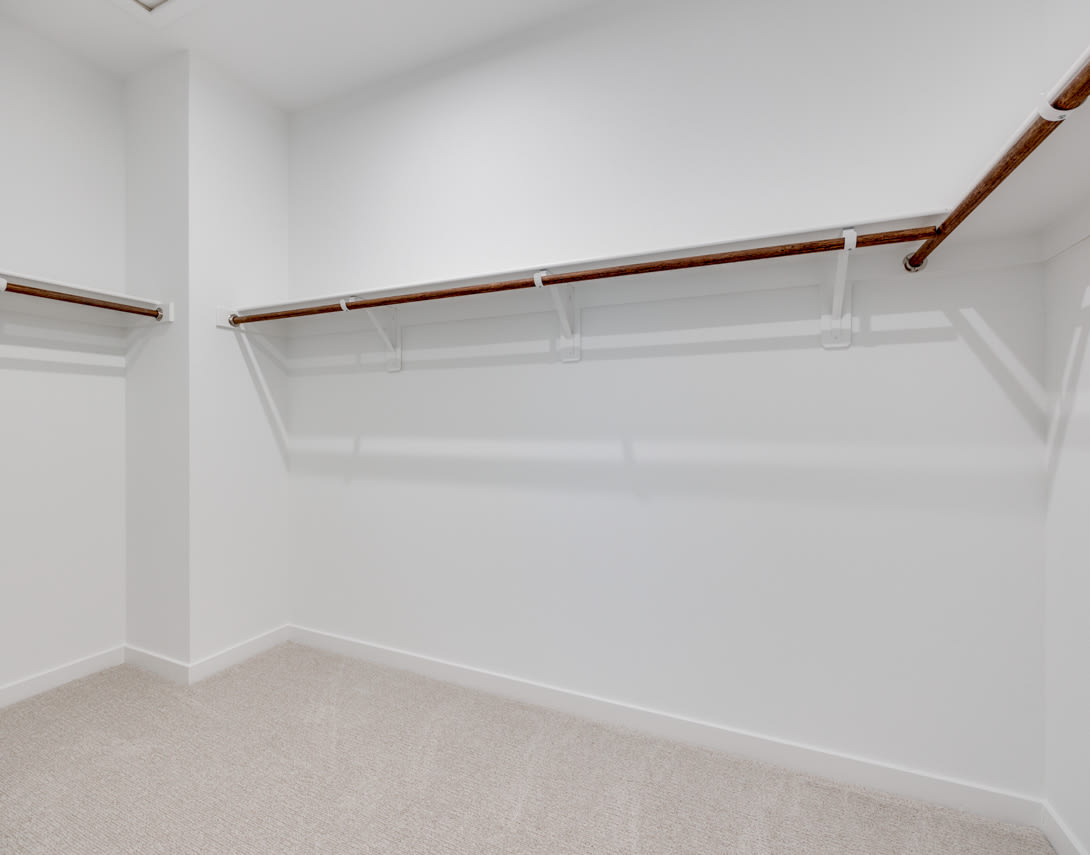 A spacious, well-lit closet with white walls, a carpeted floor, and wooden hanging rods for storing clothes.