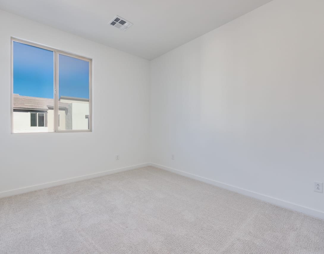 A bright, empty room with a large window overlooking a clear sky, featuring a plain white wall and a carpeted floor.