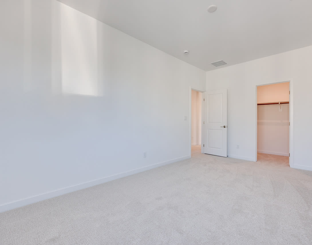 A spacious, empty room with white walls and a carpeted floor, featuring a doorway leading to another room.
