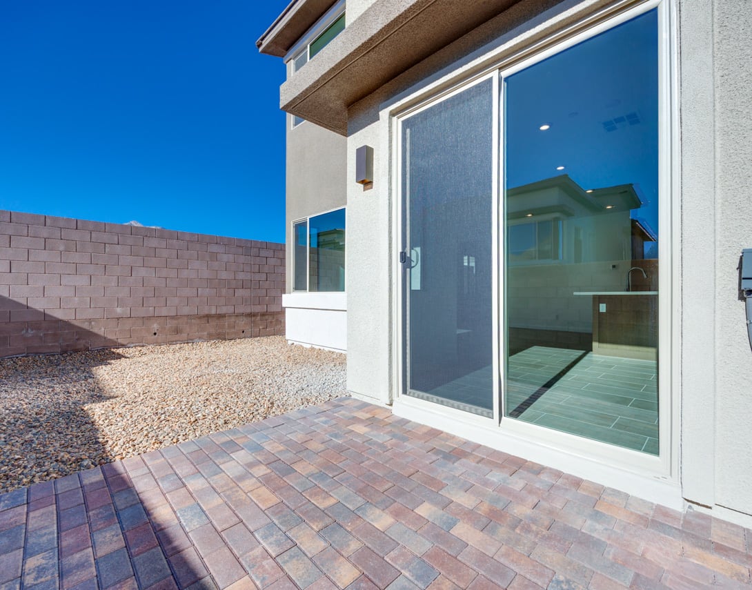 A modern, single-story house with large glass windows and doors, surrounded by a paved patio and a gravel-covered yard against a clear blue sky.
