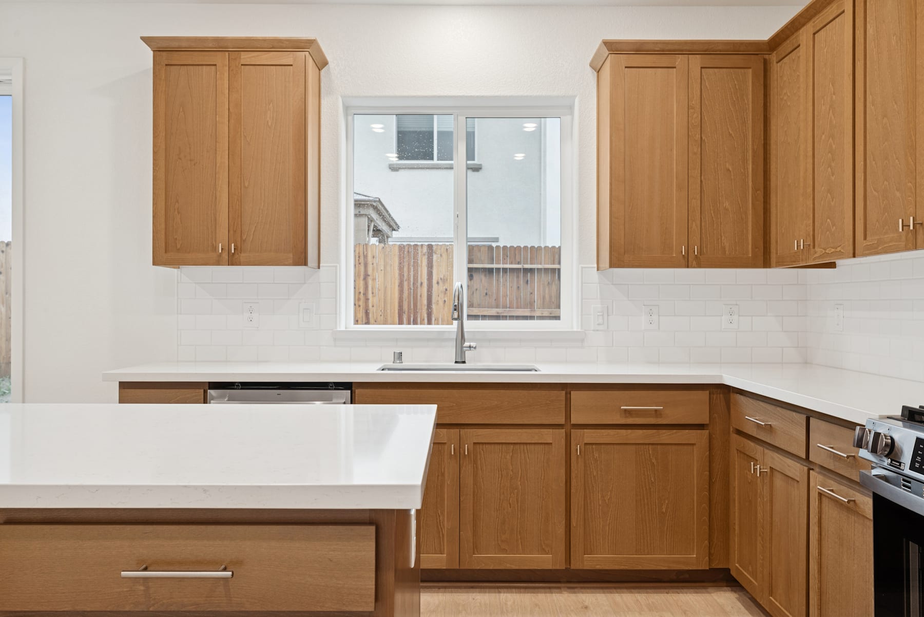 A modern kitchen with wooden cabinets, a white countertop, and a window overlooking a backyard.