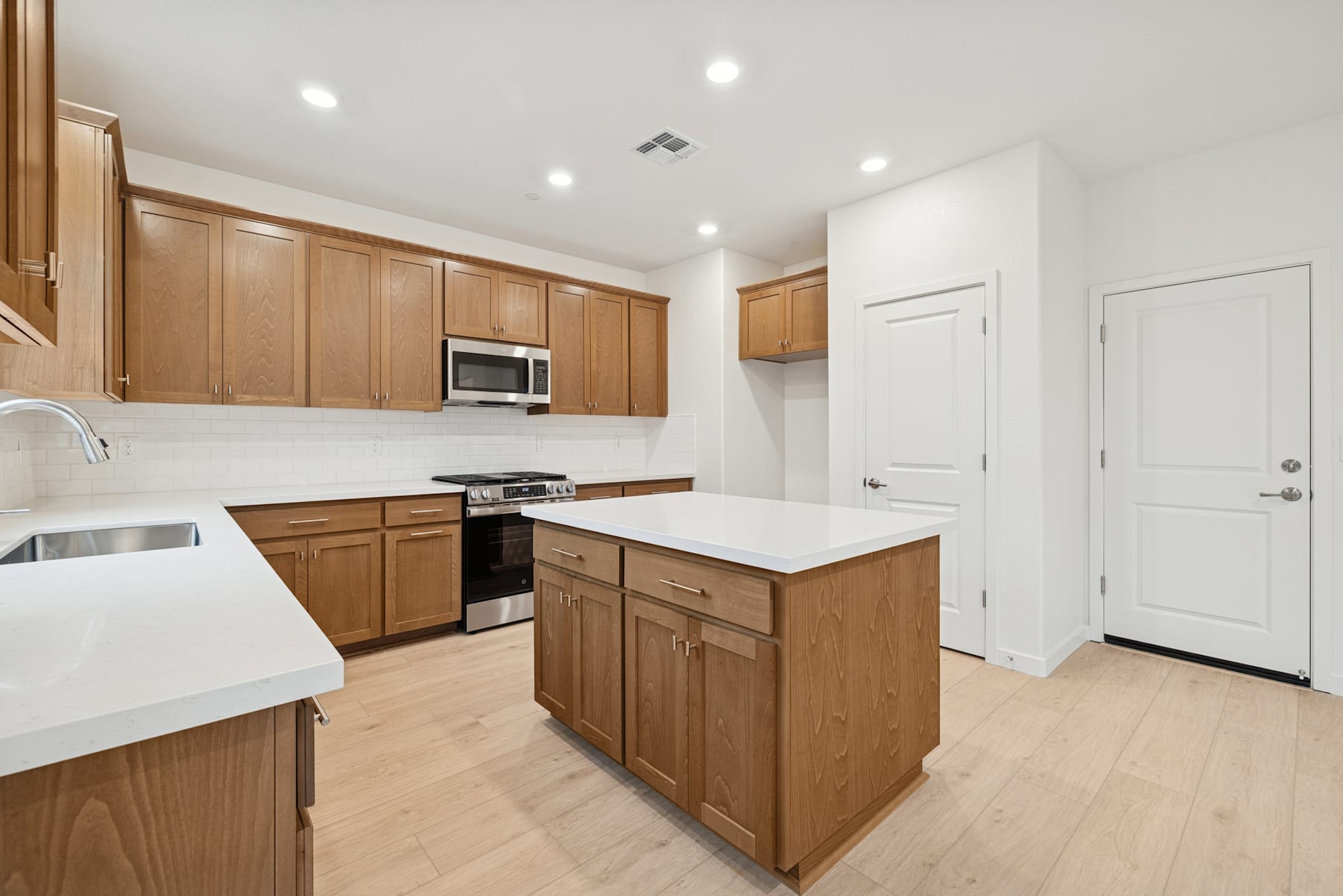 A modern, well-lit kitchen with wooden cabinets, a white countertop, and stainless steel appliances, set against a backdrop of white walls and a wooden floor.