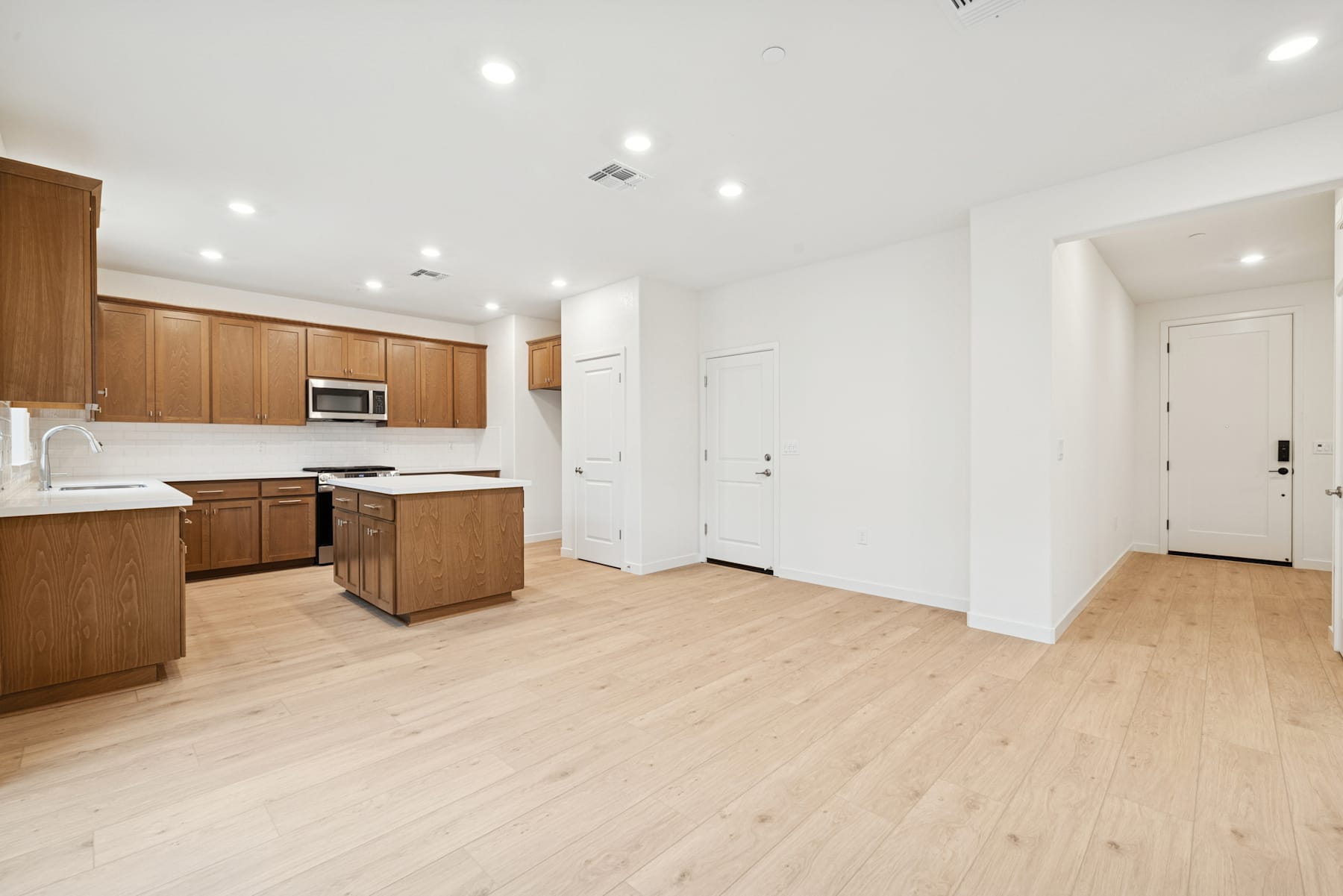 A spacious and modern kitchen with wooden cabinets, white walls, and a hardwood floor, leading into a hallway with a white door.