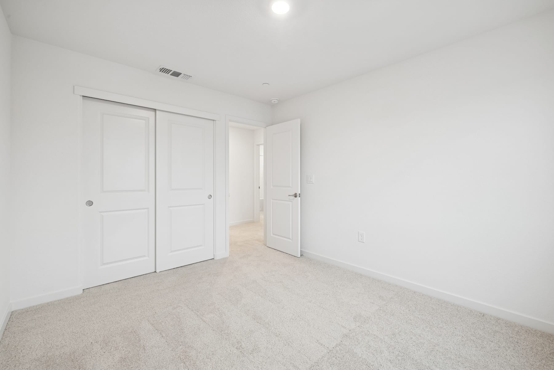 A simple, minimalist bedroom with white walls, a carpeted floor, and two closed doors in the background.