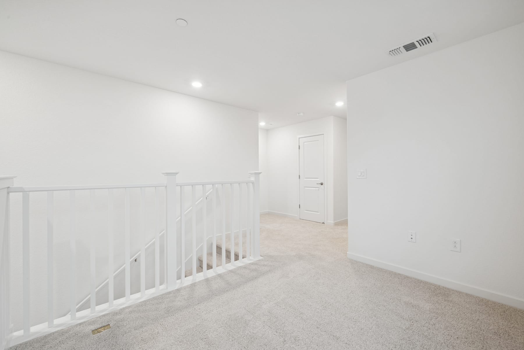 A bright, open hallway with white walls, a white railing, and a carpeted floor leading to a closed door.