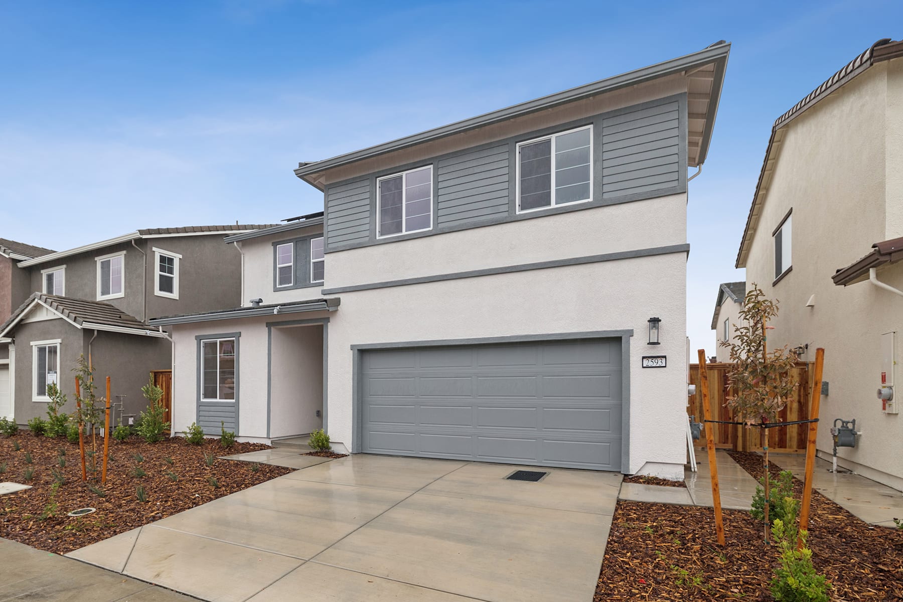 A modern two-story residential building with a gray exterior, a garage door, and a concrete walkway leading to the entrance, set against a clear blue sky.