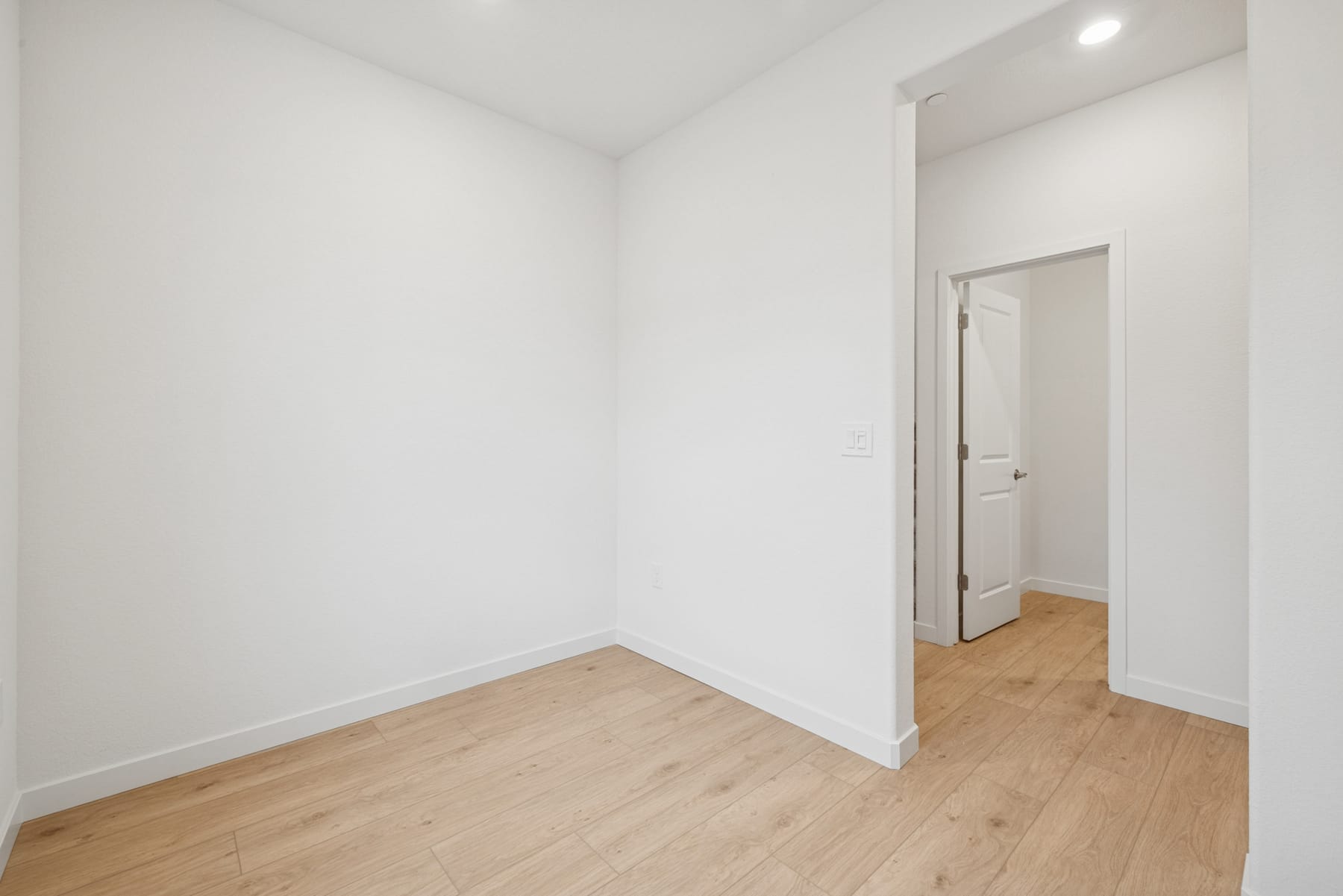 A bright, minimalist room with white walls and a wooden floor, featuring an open doorway leading to another room.
