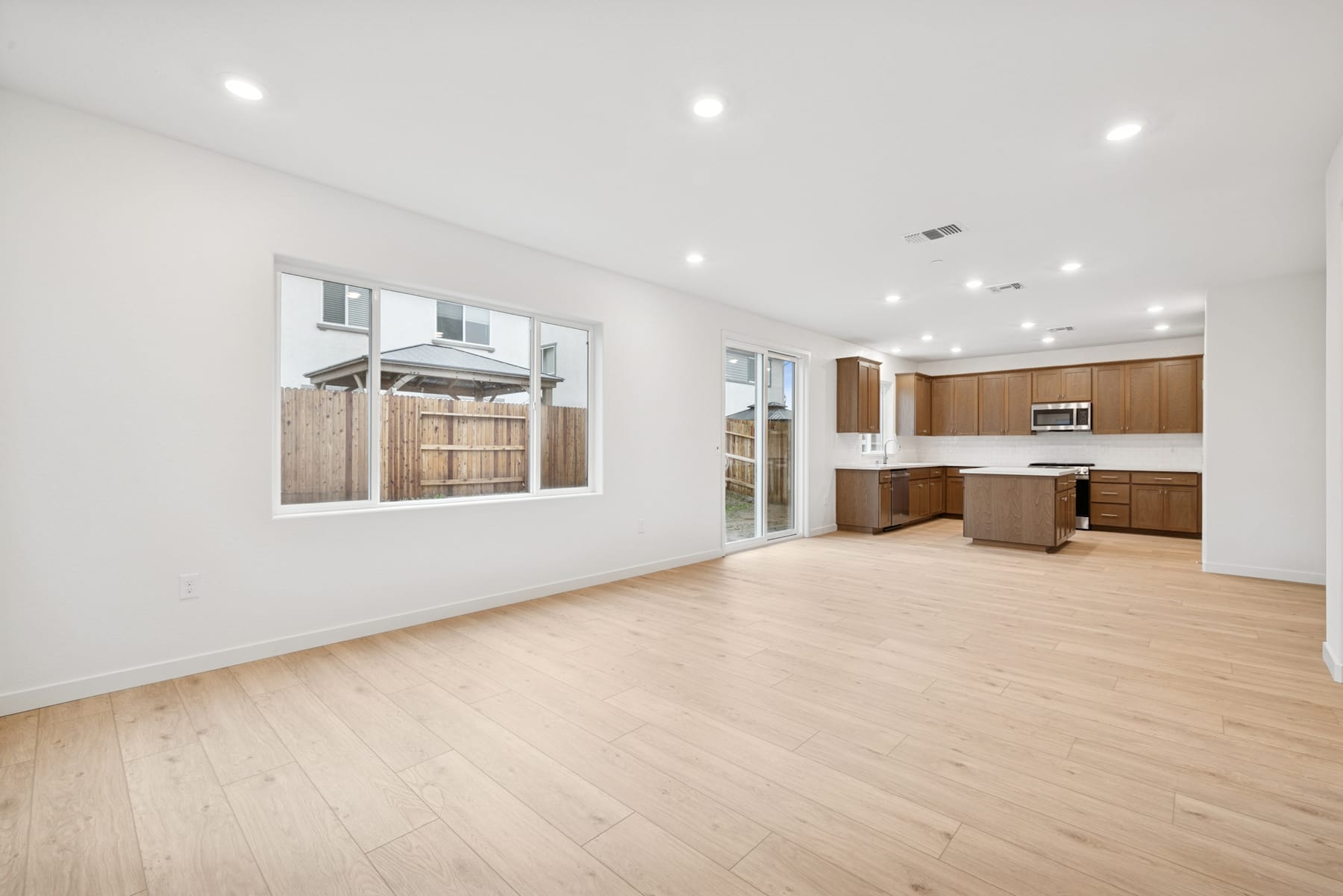 A spacious, open-concept living area with a kitchen visible in the background, featuring light-colored hardwood floors, white walls, and recessed lighting.