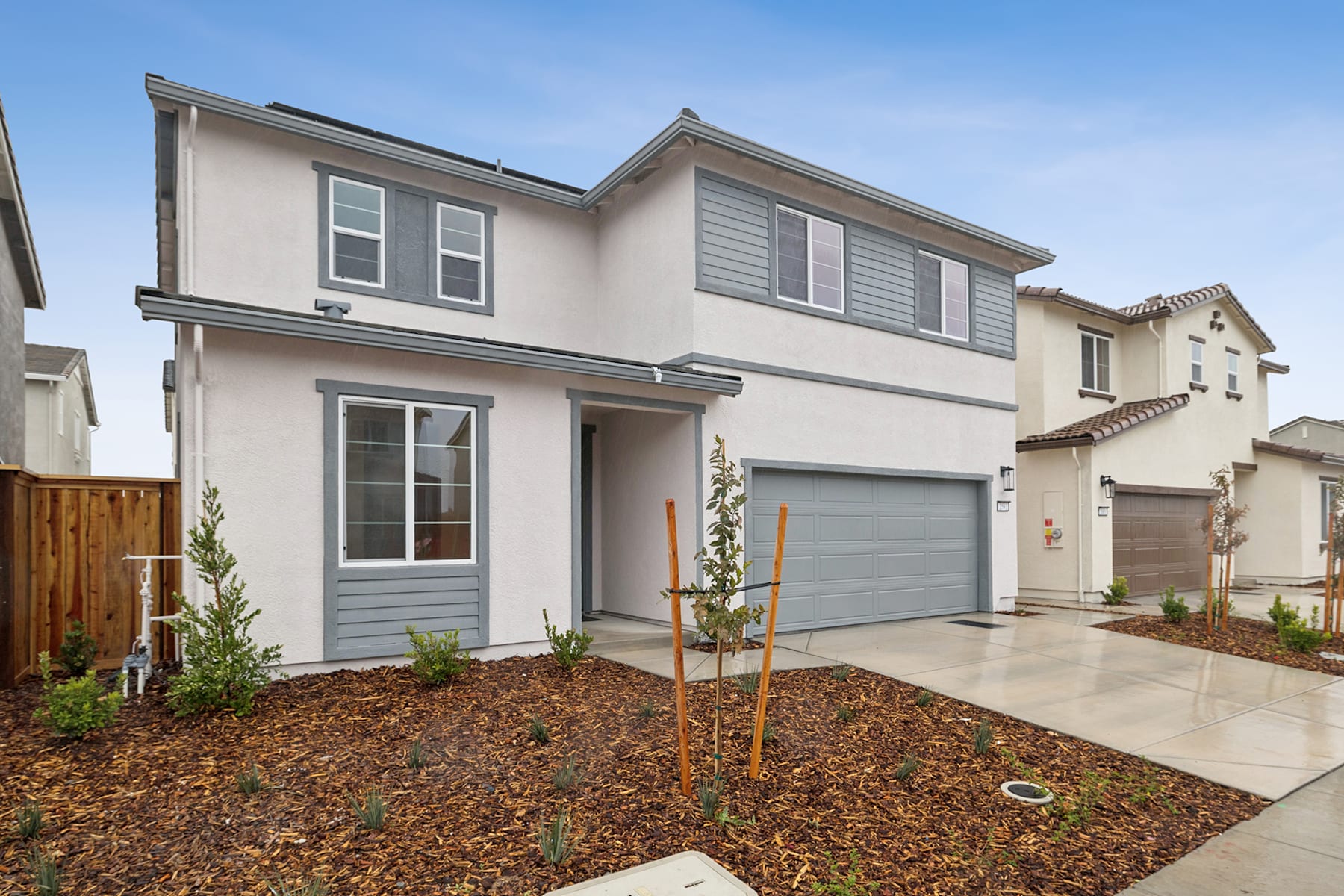 A two-story residential house with a gray exterior, a garage door, and a landscaped front yard with mulch and small plants.