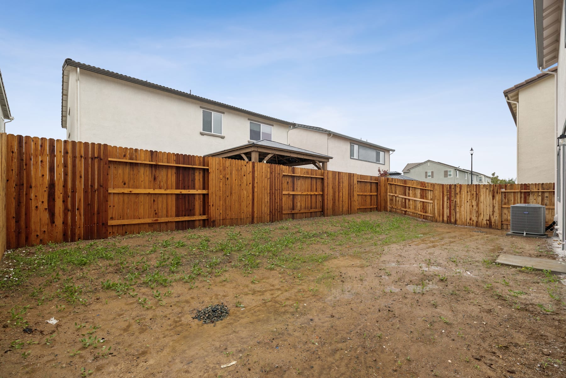 A fenced backyard with a wooden fence, a grassy area, and residential buildings in the background under a clear blue sky.