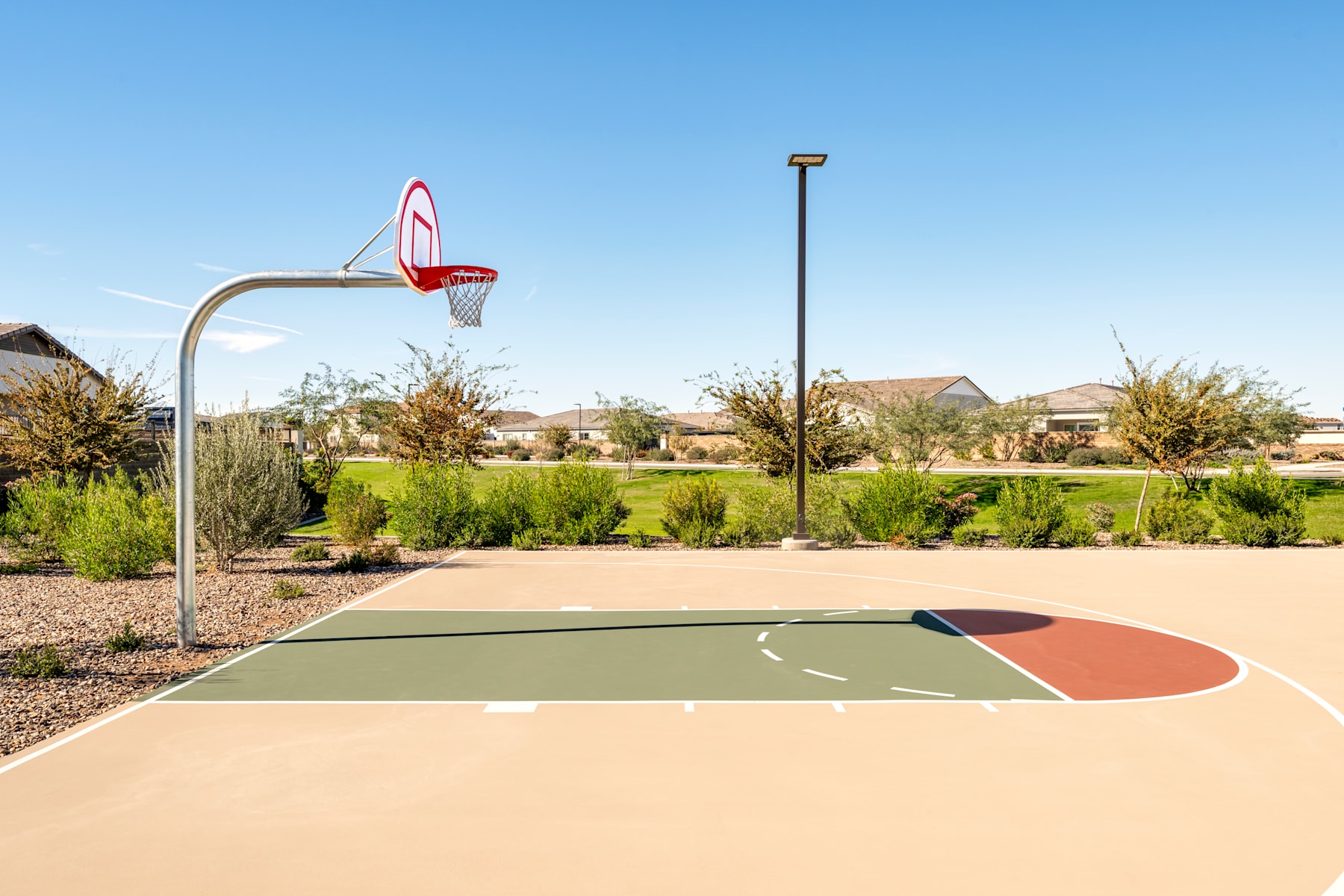 A basketball court with a hoop and backboard stands in the foreground, surrounded by a well-maintained landscaped area with houses visible in the background.