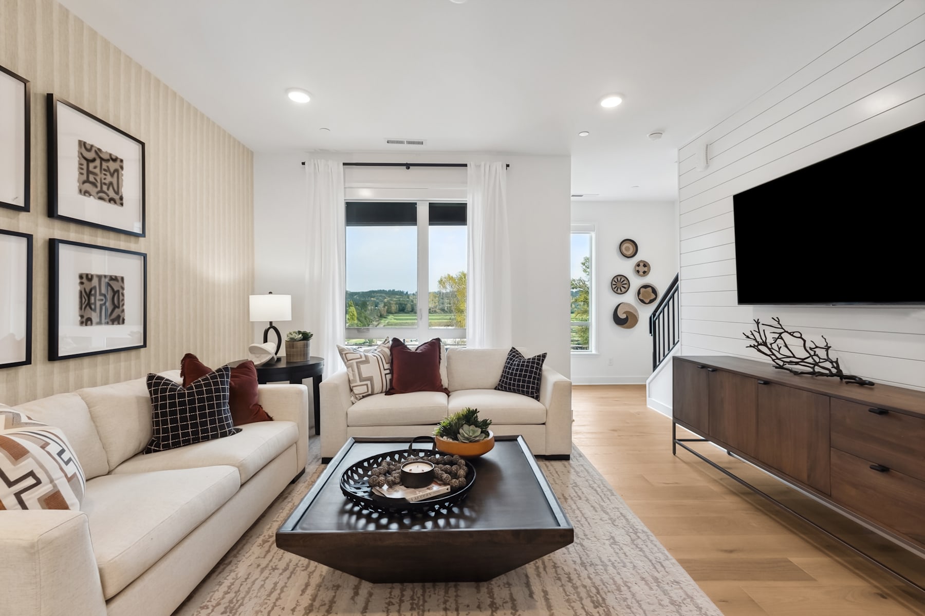 A cozy and modern living room with a large sofa, a wooden coffee table, and framed artwork on the walls, overlooking a scenic outdoor view through the large window.