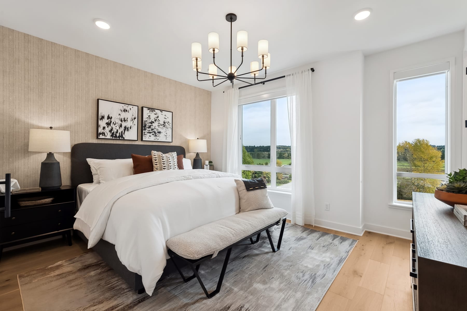 A spacious and well-lit bedroom with a large window, a modern chandelier, and a cozy bed with white bedding and decorative pillows. The room features wooden floors and neutral-toned walls, creating a warm and inviting atmosphere.