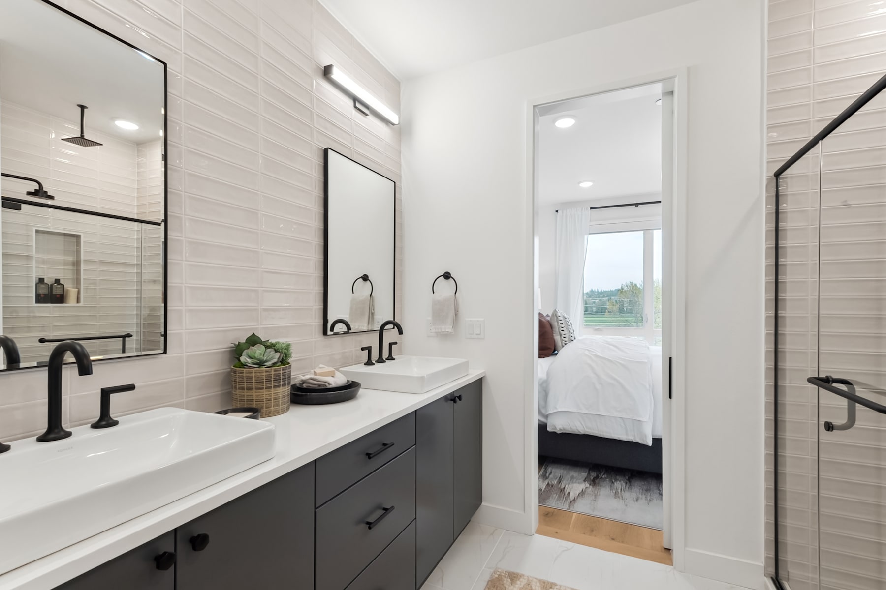 A modern and minimalist bathroom with a double vanity, black fixtures, and a view of a bedroom through an open doorway.