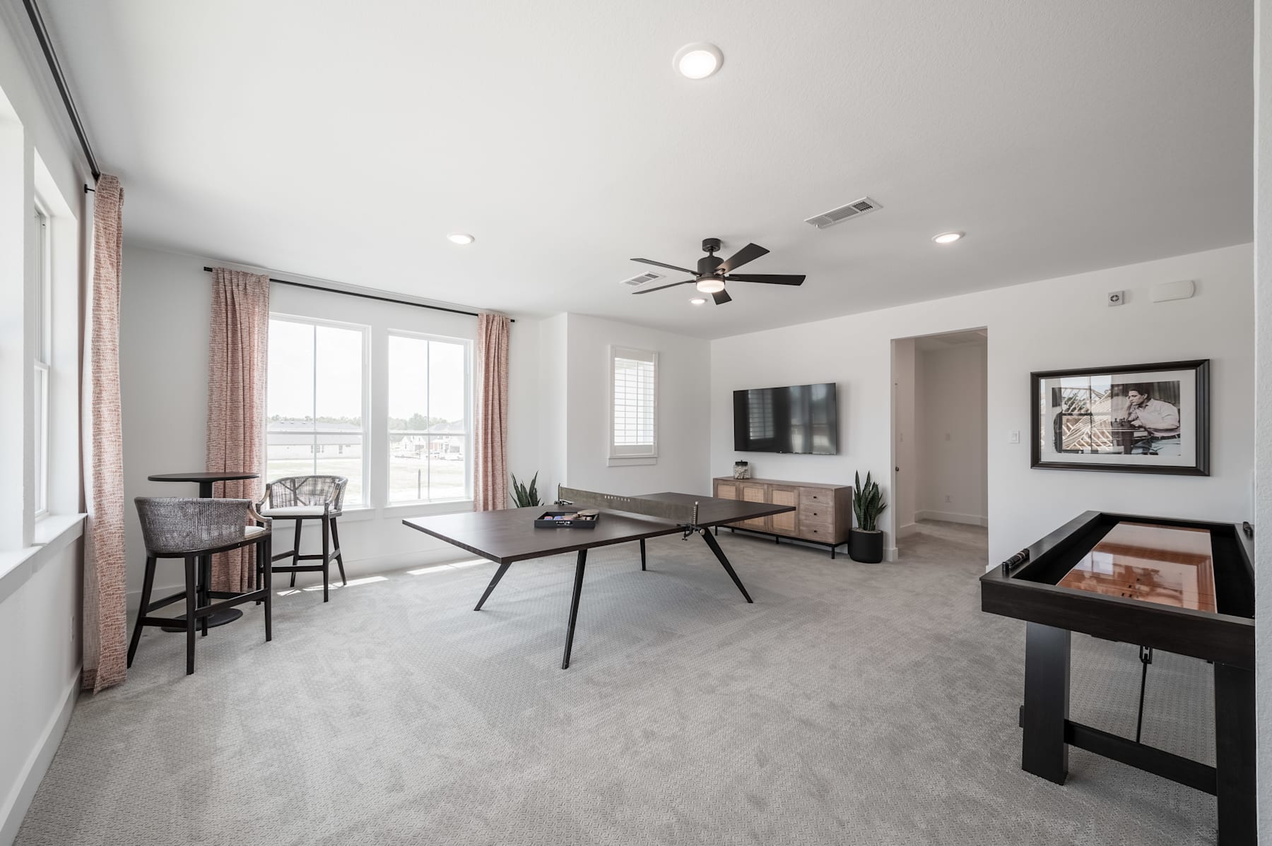 A modern and minimalist living room with a large window, a ceiling fan, and various furniture pieces such as a coffee table, a TV stand, and bar stools.