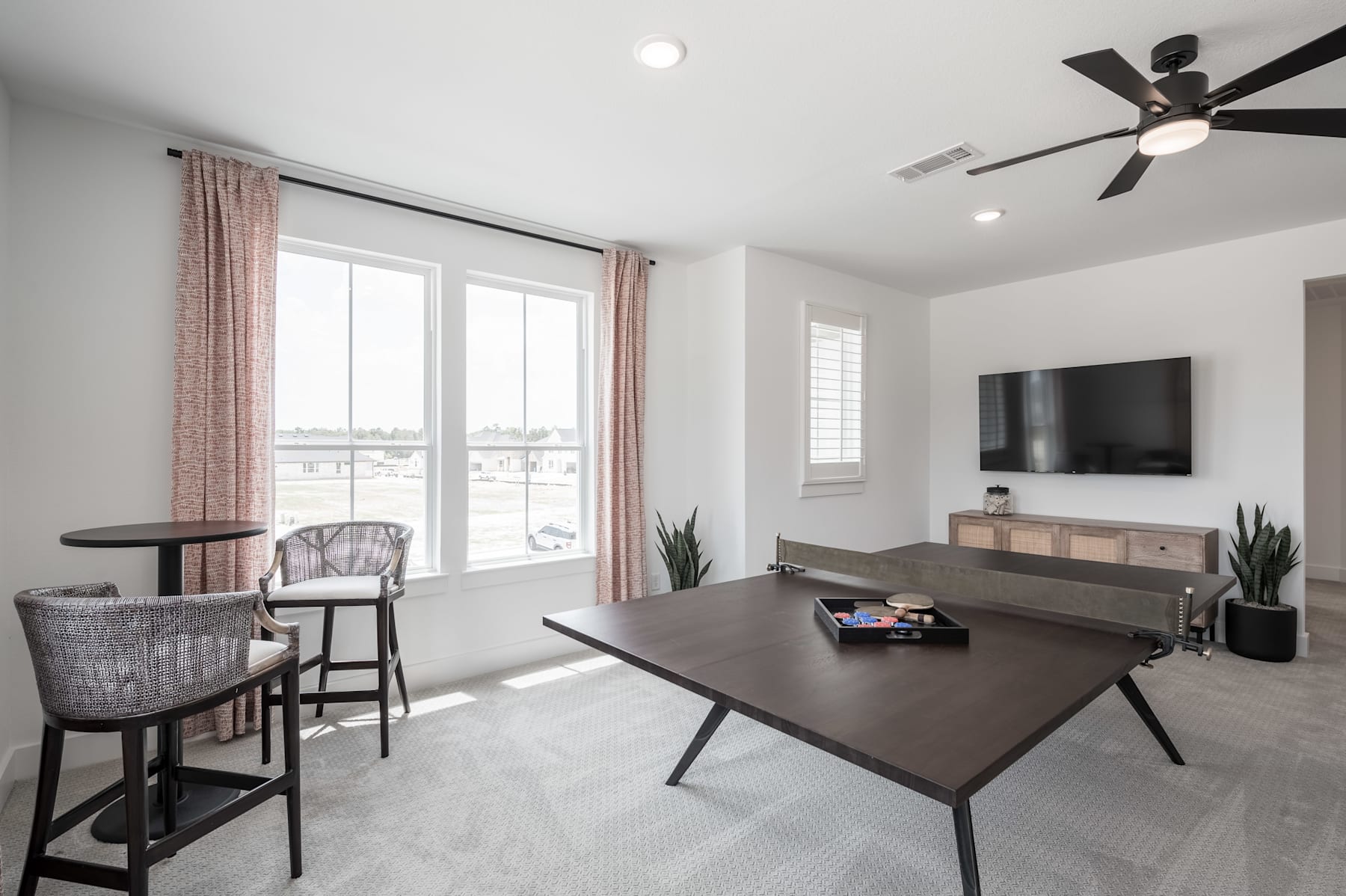 A modern and minimalist living room with a large window, a ceiling fan, and a wooden table with chairs in the foreground.