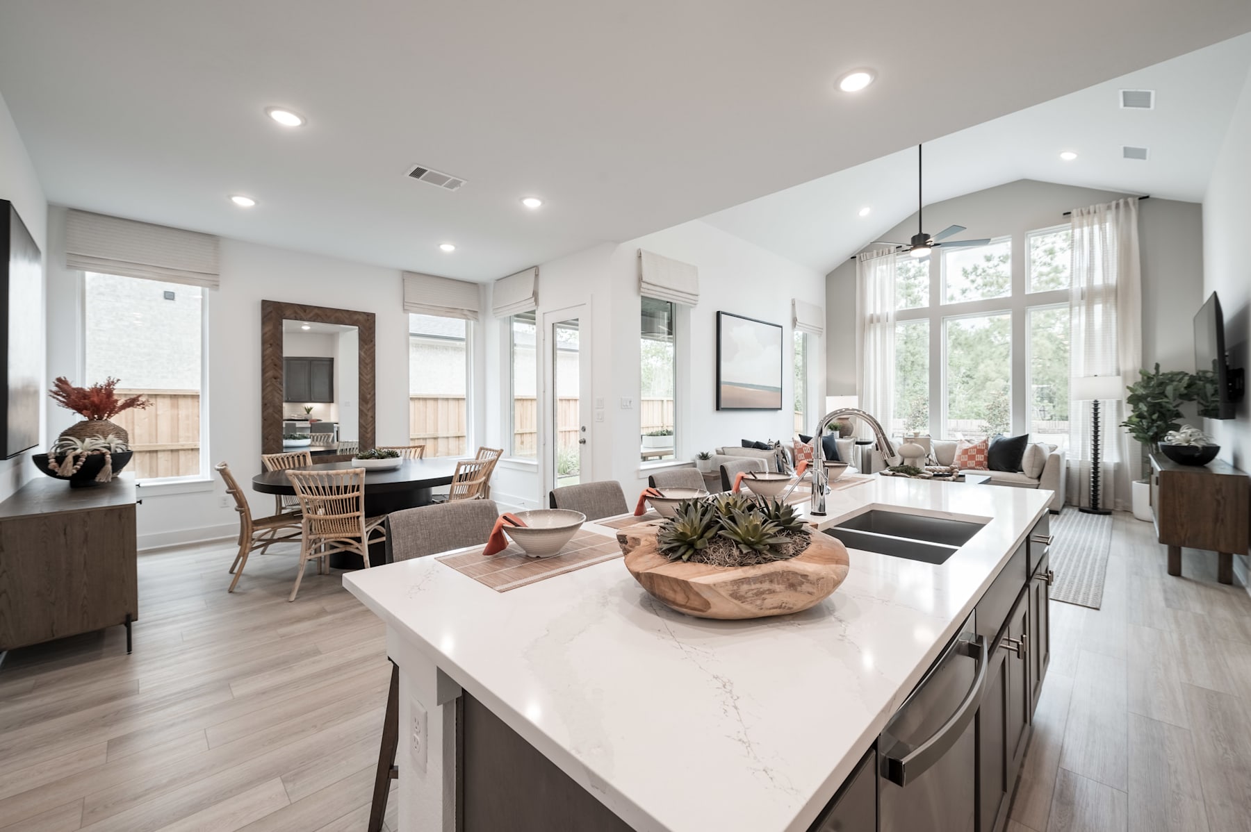 A modern, open-concept living space with a white kitchen island, wooden furniture, and large windows allowing natural light to flood the room.
