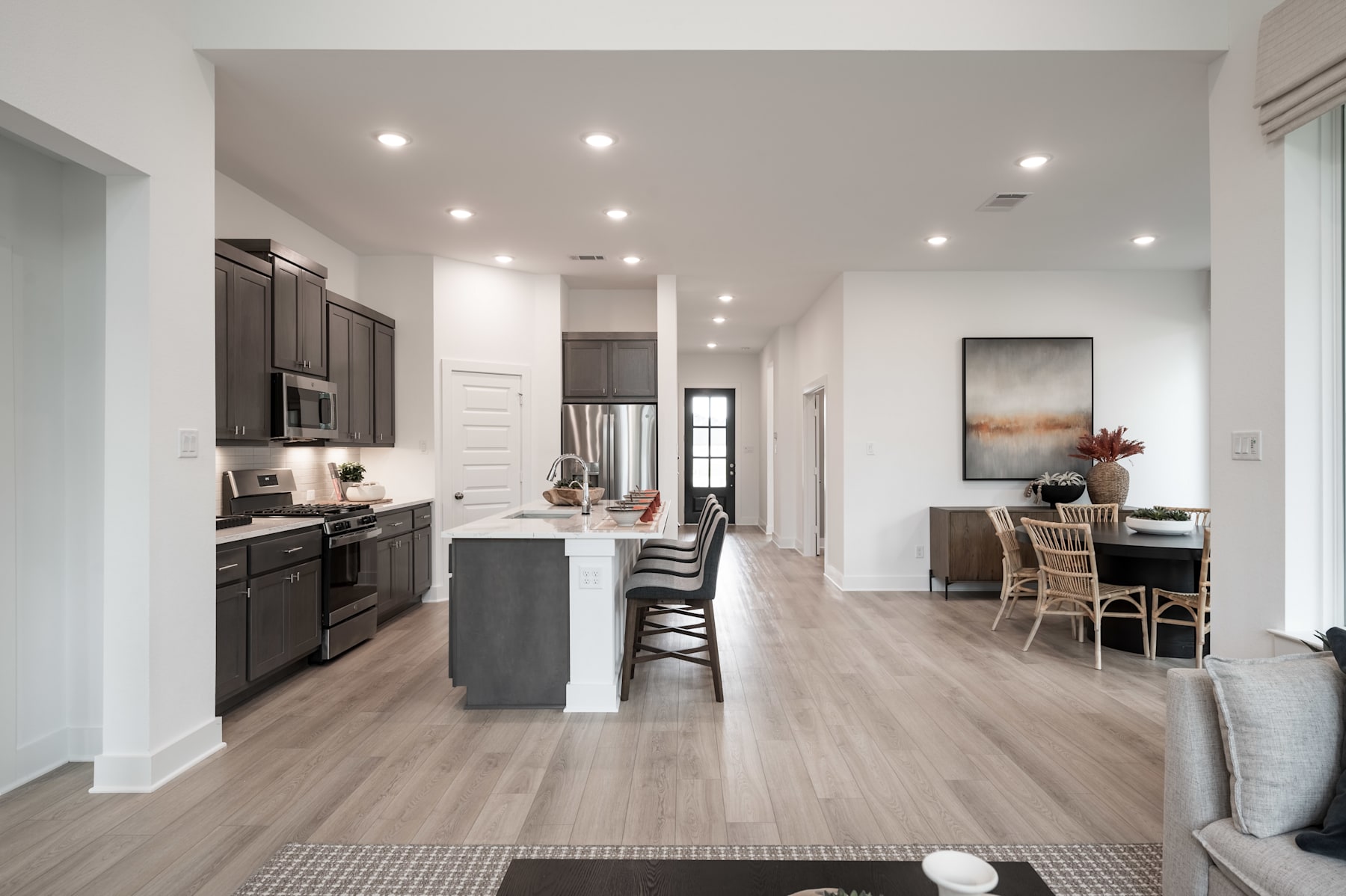 A modern, open-concept kitchen and living area with dark cabinets, light hardwood floors, and a cozy seating area in the background.