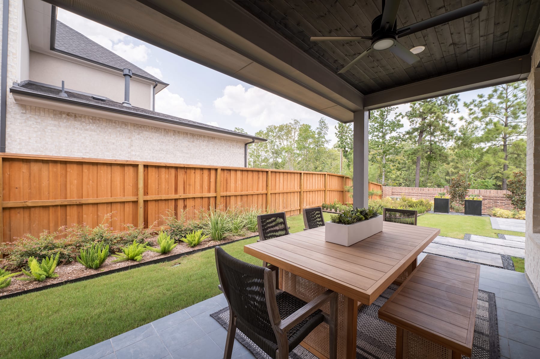 A cozy outdoor patio with a wooden table and chairs, surrounded by a lush green lawn and a wooden fence, with a covered roof overhead and trees in the background.