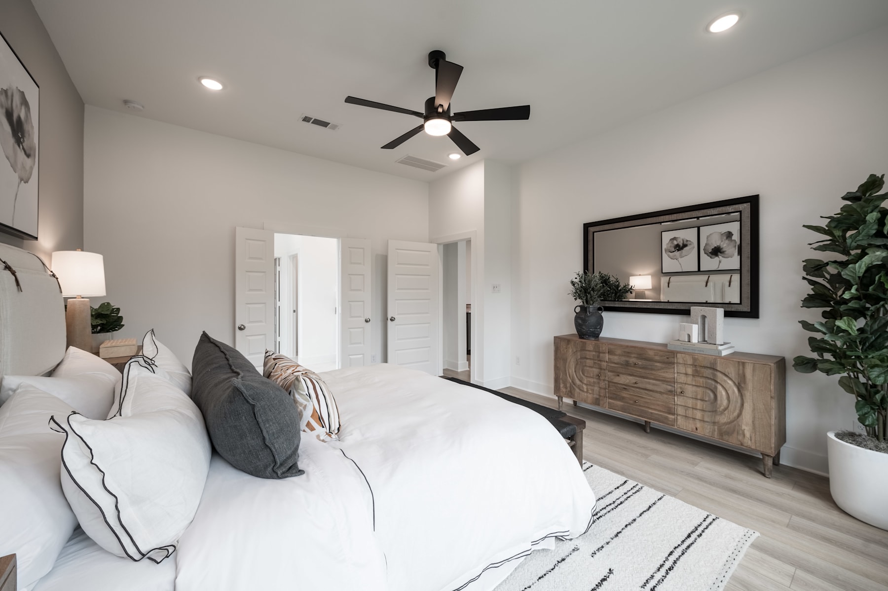 A cozy and modern bedroom with a white bed, gray and white pillows, a wooden dresser, and a ceiling fan, set against a neutral-toned wall with framed artwork.