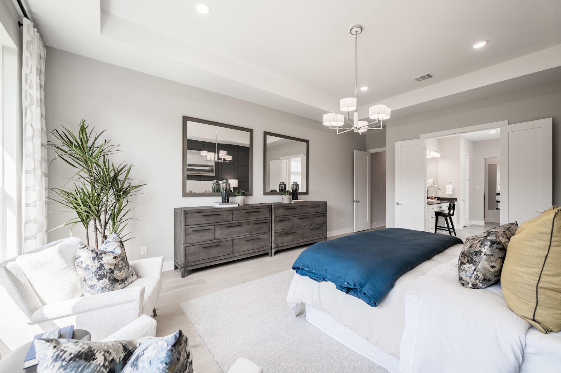A spacious and modern bedroom with a large bed, a dresser, and a chandelier overhead, surrounded by neutral-toned walls and decor.