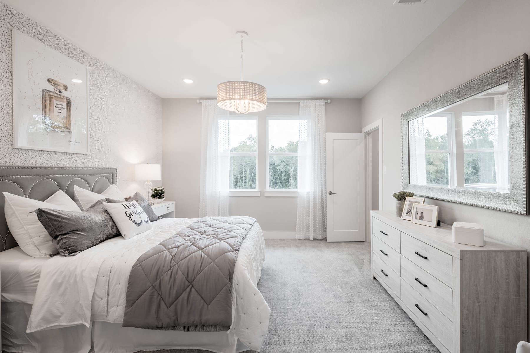 A spacious and elegantly designed bedroom with a plush gray upholstered headboard, a large window, and a dresser with drawers, all in a serene and calming white and gray color scheme.