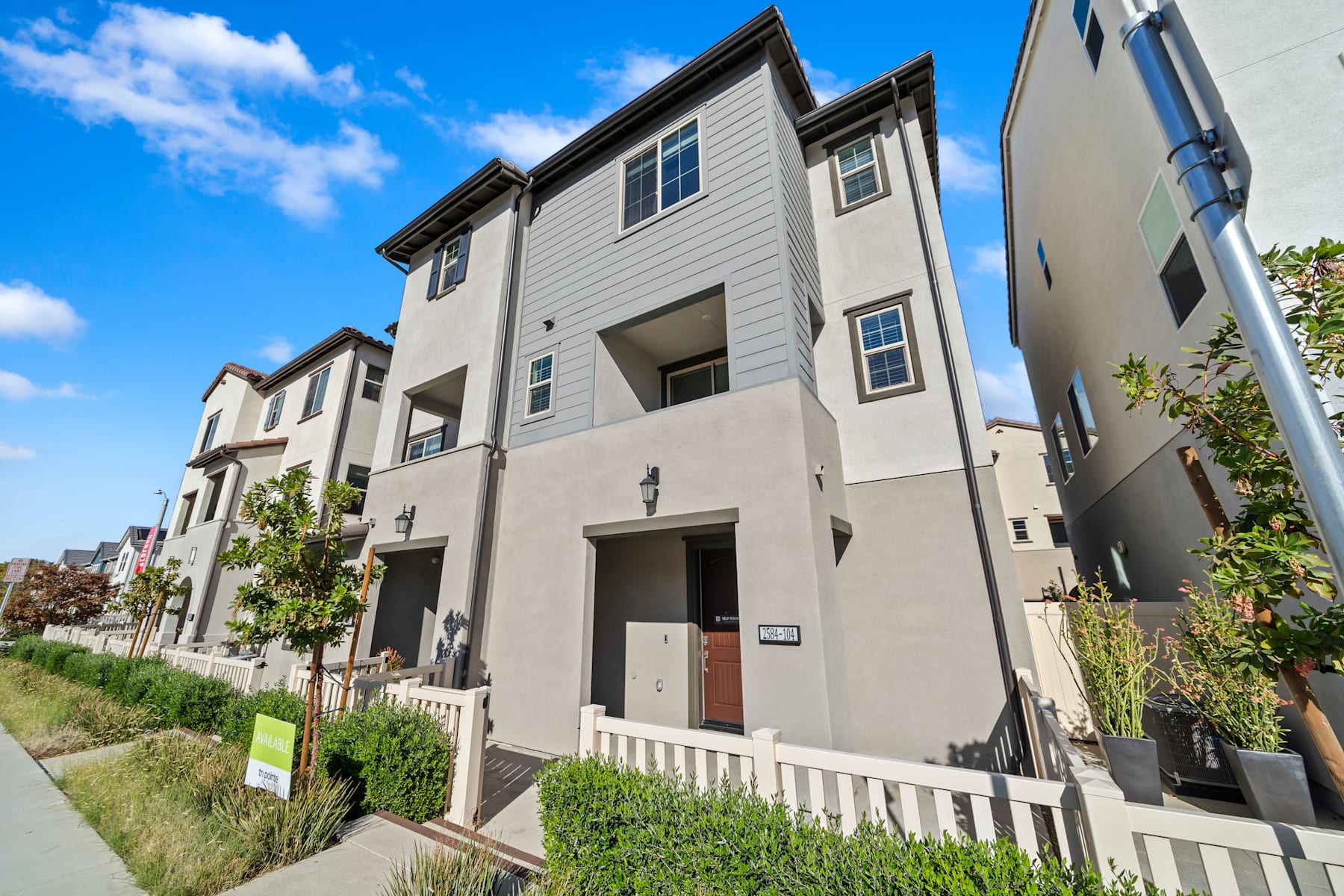 The image depicts a row of modern, multi-story townhouses with gray siding, white trim, and red doors, set against a backdrop of a clear blue sky with scattered clouds.