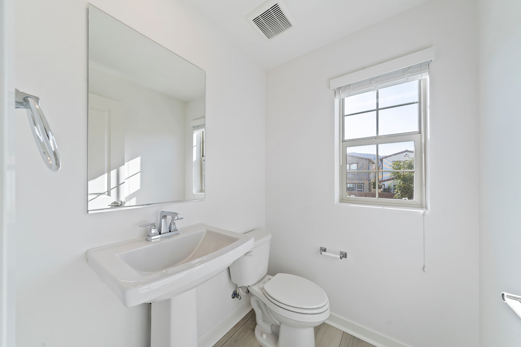 A bright and airy bathroom with a white sink, toilet, and large window overlooking a residential neighborhood.