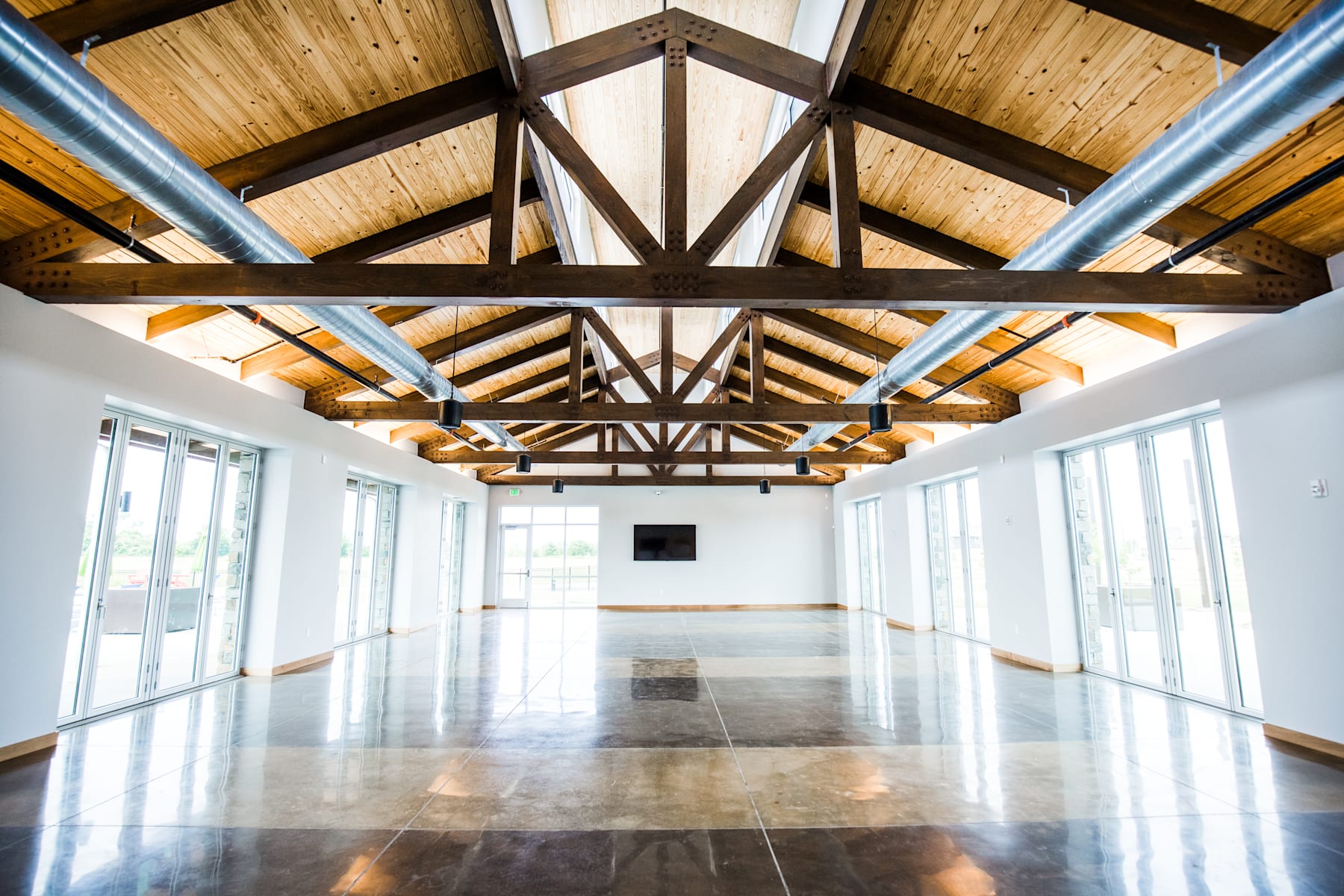 A spacious and well-lit interior with a high, wooden-beamed ceiling, polished hardwood floors, and large windows allowing natural light to flood the room.