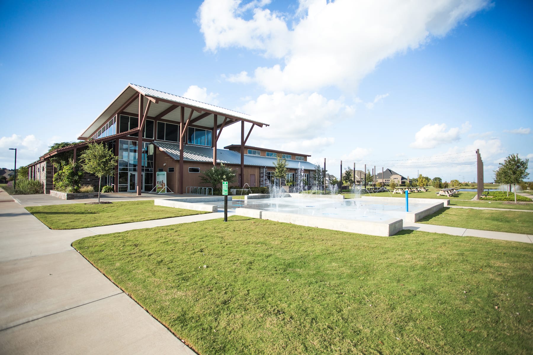 A modern, multi-story building with large windows and a covered porch stands in a grassy area with a water fountain feature in the foreground, against a backdrop of a clear blue sky with fluffy white clouds.