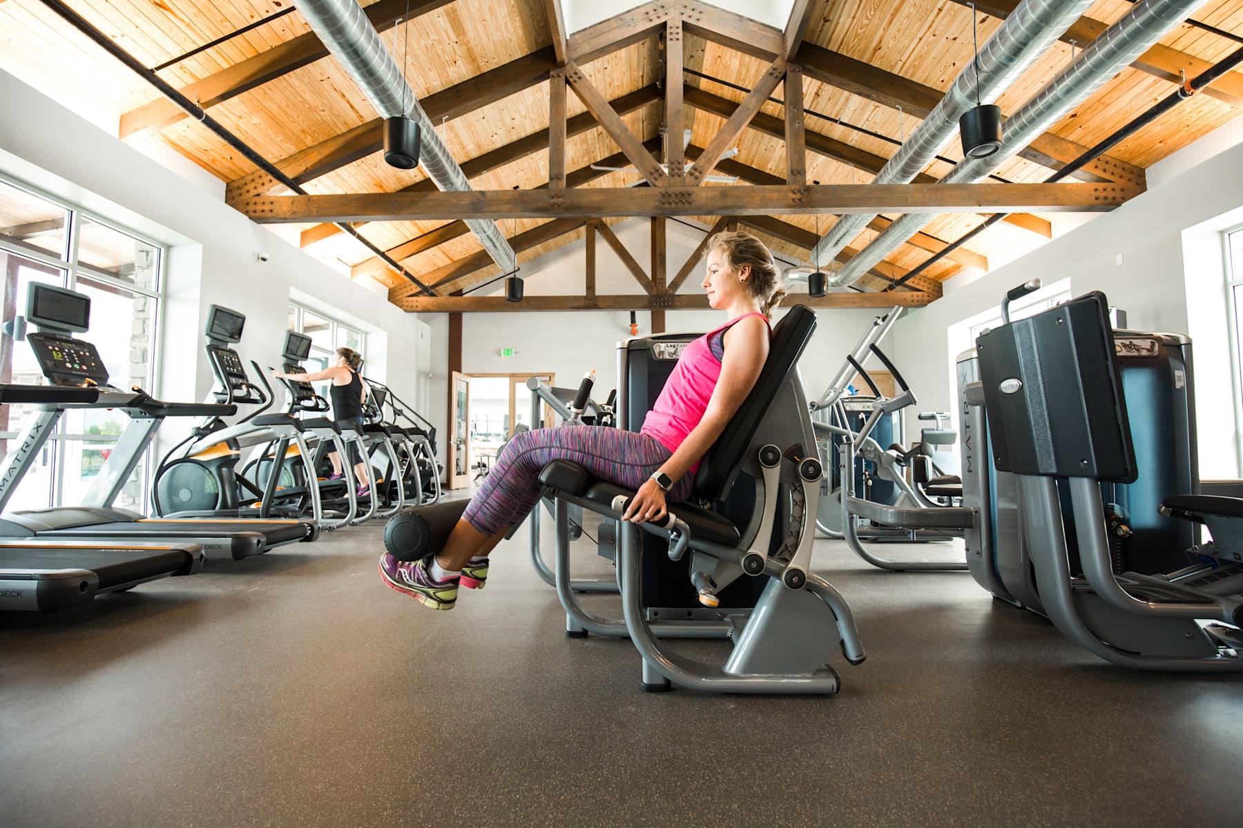 A woman in a pink top and leggings is exercising on a machine in a well-equipped gym with wooden beams and high ceilings.
