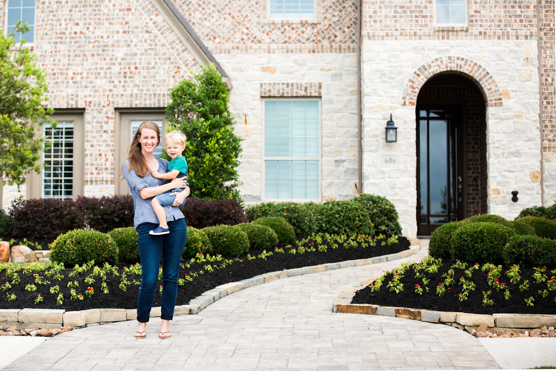 A woman holding a young child stands in front of a large, brick building with an arched entryway, surrounded by neatly trimmed bushes and a paved walkway.