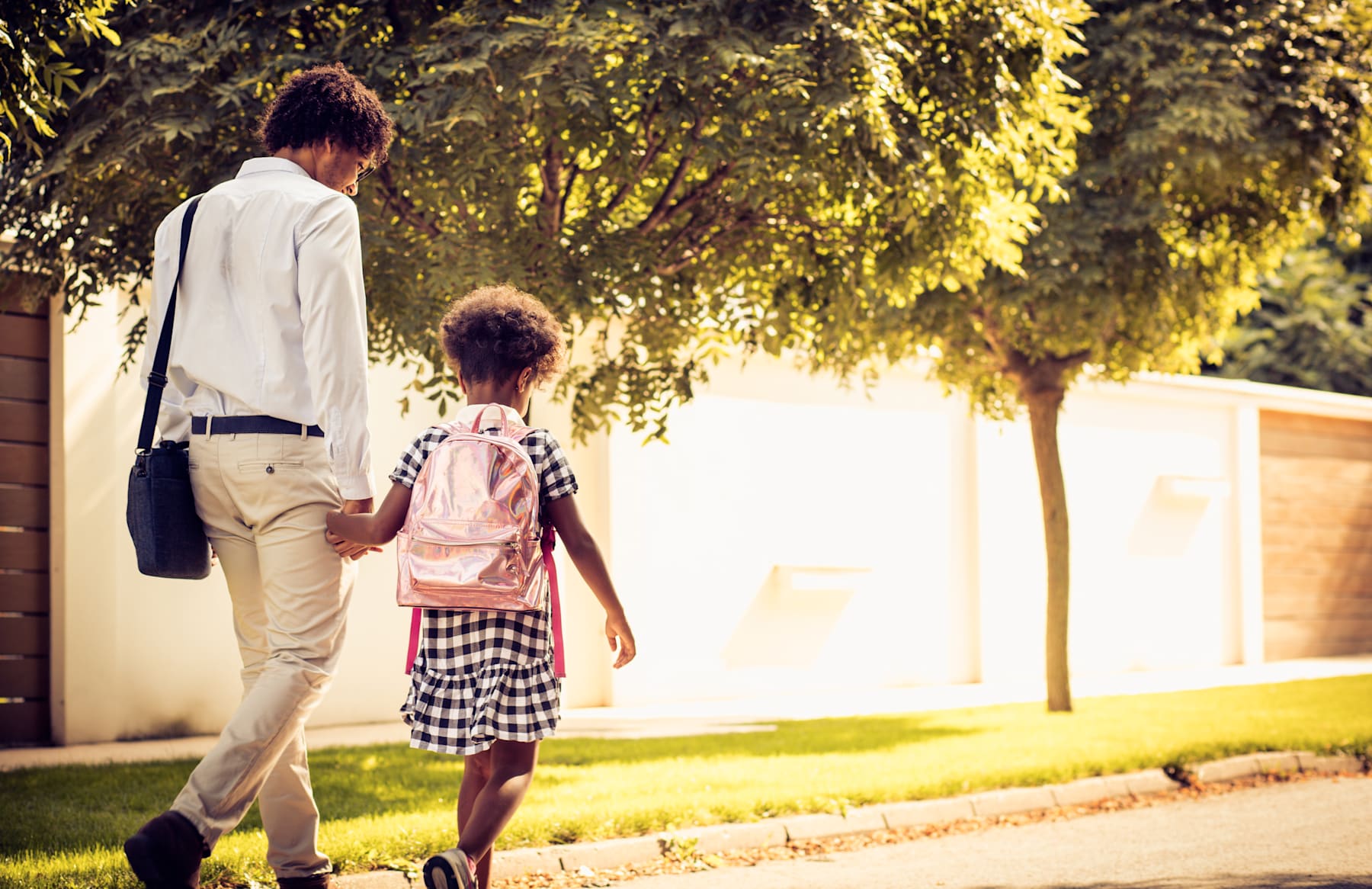 A person in a light-colored jacket is walking alongside a young child in a checkered dress, against a backdrop of lush greenery and a sunny outdoor setting.