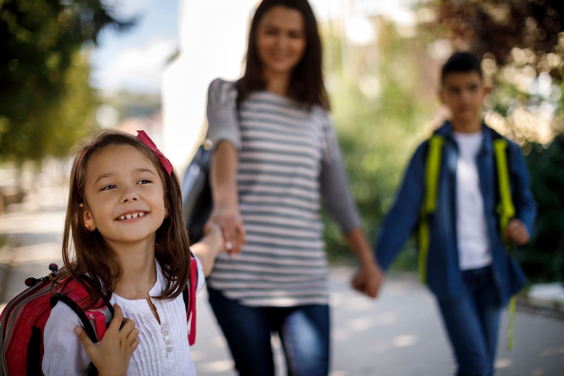 A young girl with a red backpack is smiling in the foreground, while two older individuals, likely her parents, are walking behind her in a park-like setting.