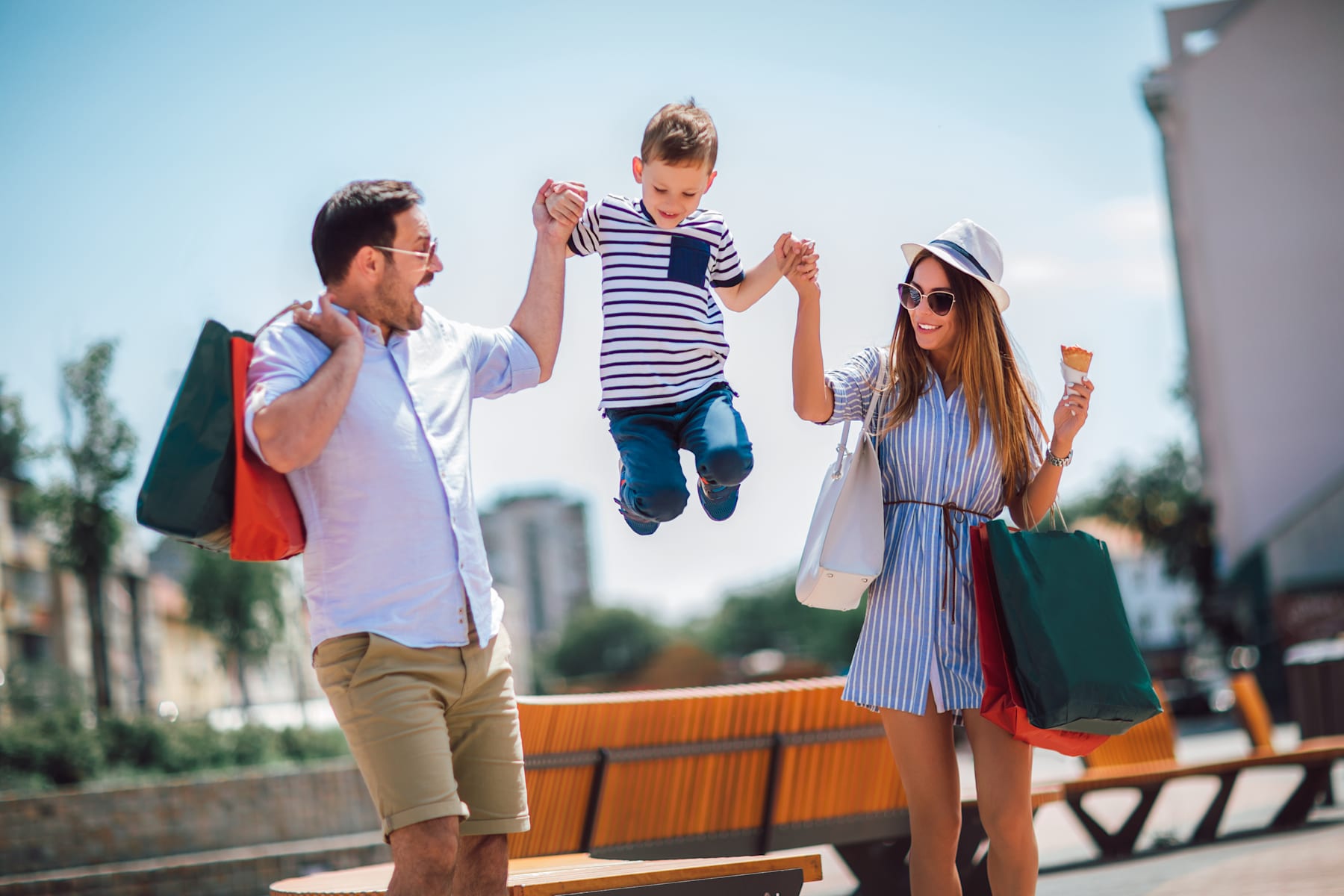 A group of young people, a man and two women, are walking together and one of the women is jumping up in the air, holding shopping bags, against a backdrop of buildings and a sunny outdoor setting.