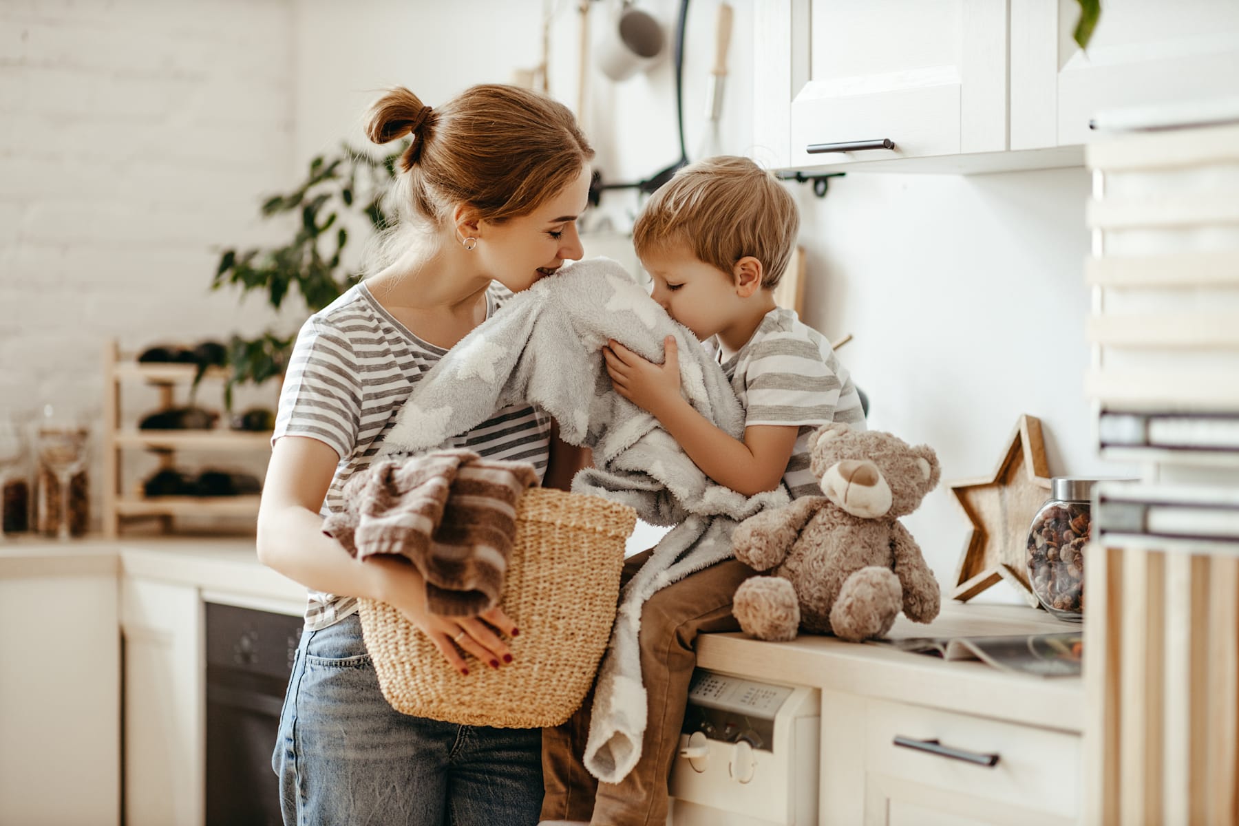A young woman is embracing a young boy, both holding stuffed animals, in a cozy, home-like setting with shelves and decor visible in the background.