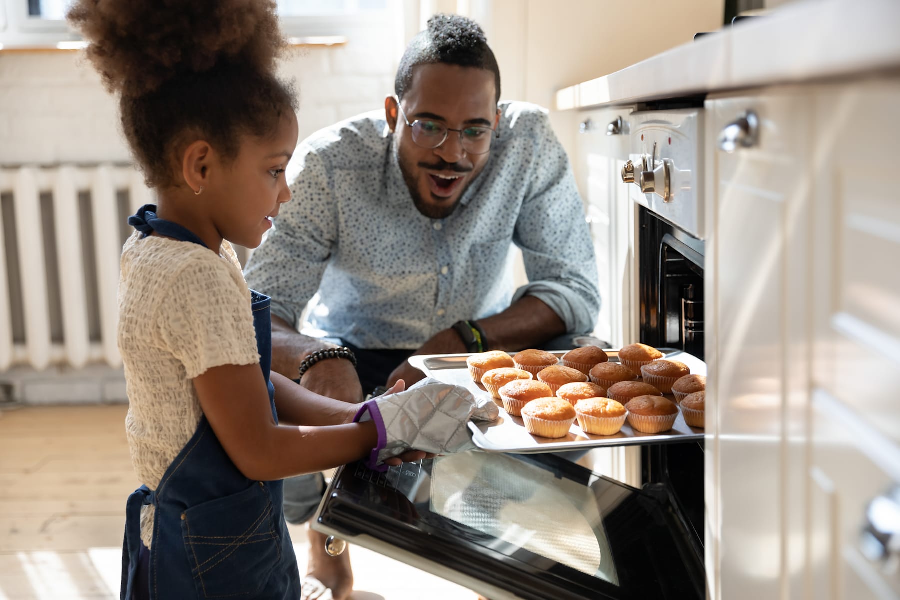 A man in a gray sweater is smiling and holding a tray of cupcakes as he interacts with a young girl in the kitchen.