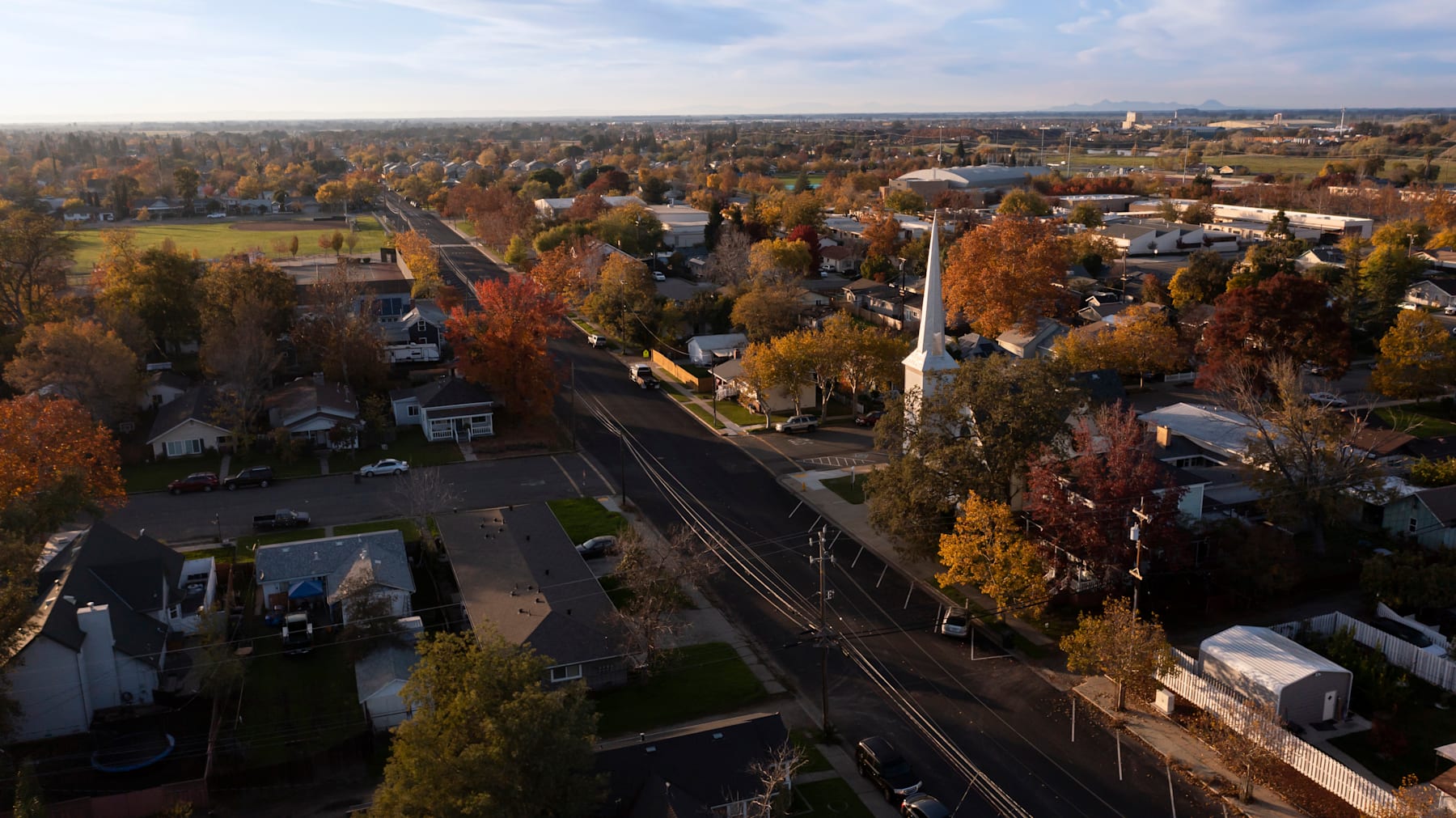 A scenic aerial view of a residential neighborhood in autumn, with colorful foliage lining the streets and railroad tracks running through the center.