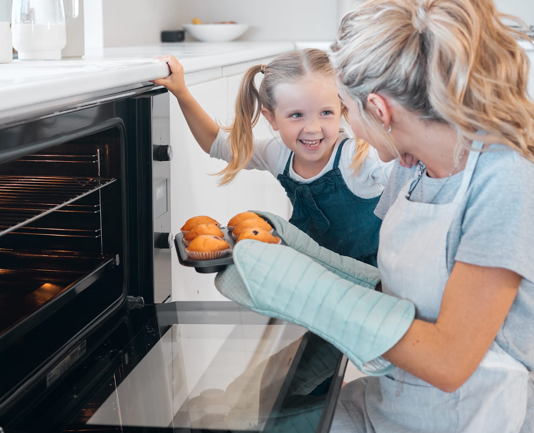 A young girl and her mother are baking together, with the mother helping the daughter check on the food in the oven.