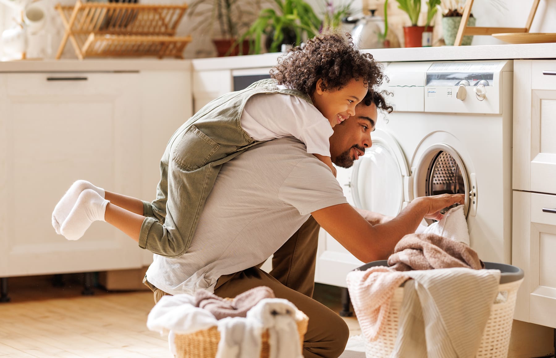 A man with curly hair is crouched down in front of a washing machine, sorting through a pile of laundry in a cozy, plant-filled home environment.