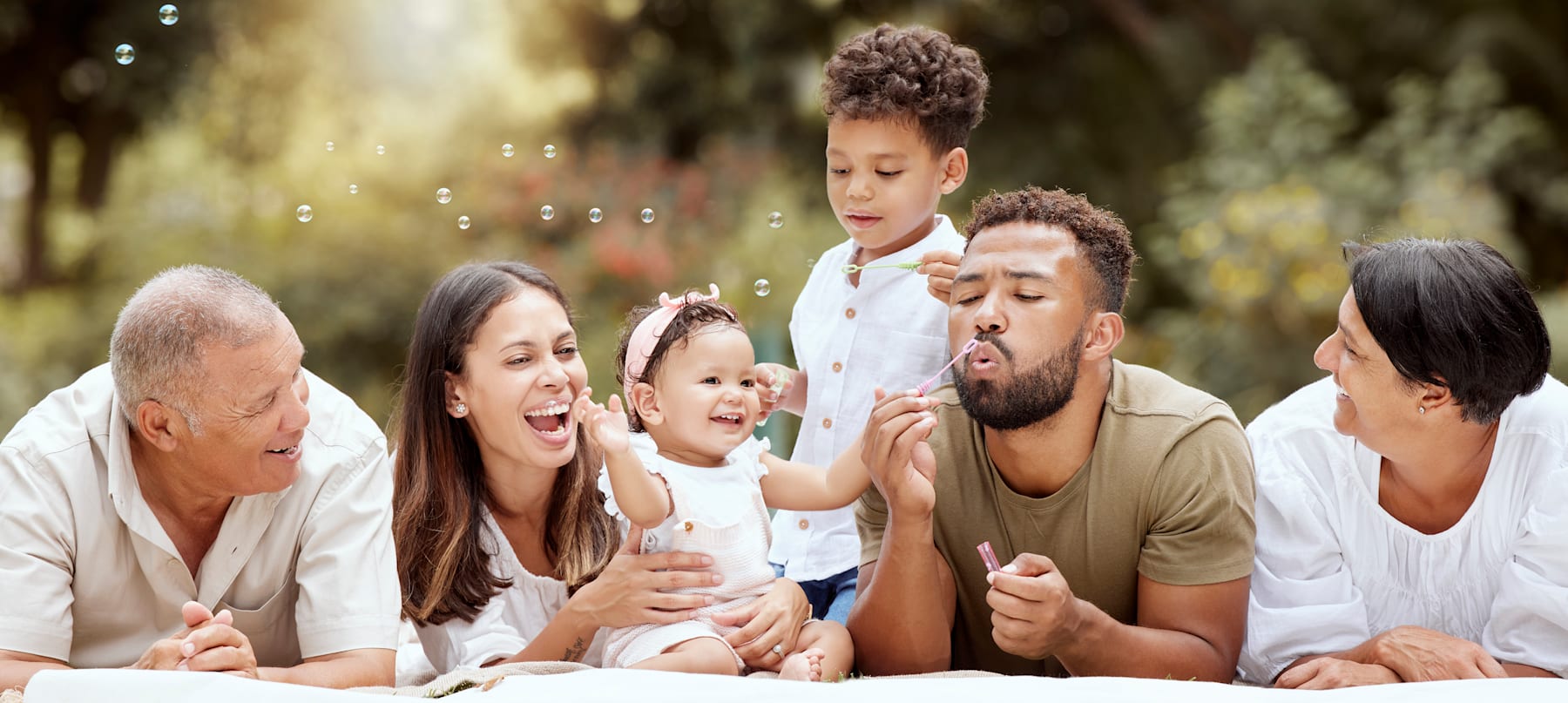 A happy, diverse family sitting together outdoors, surrounded by a blurred natural setting with sunlight filtering through.