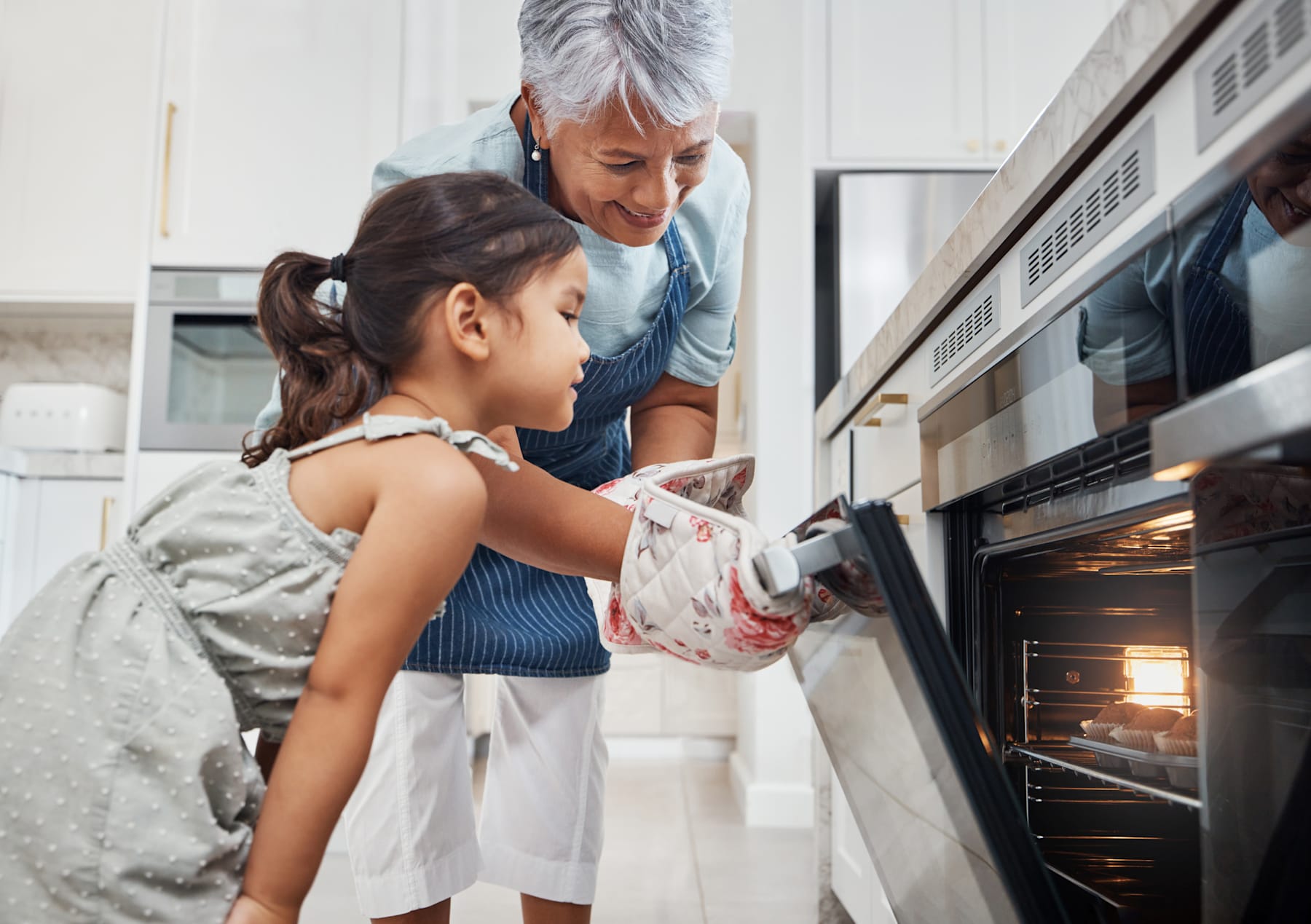 A young girl in a floral dress is standing in a kitchen, with an older adult, likely a parent or guardian, assisting her as she opens the oven door.