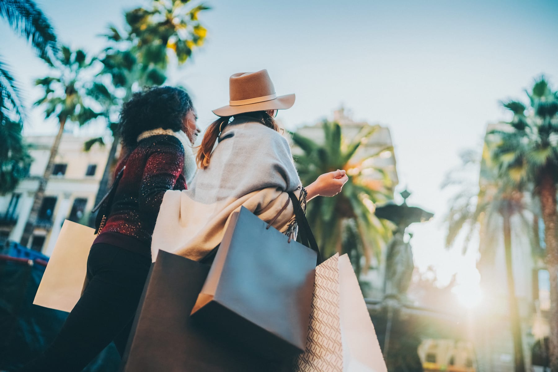 Two people, a man and a woman, are walking together while carrying shopping bags in a sunny, palm tree-lined street.