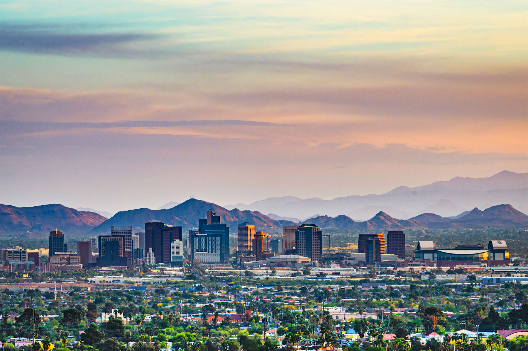A vibrant cityscape nestled among majestic mountains, with a colorful sunset sky casting a warm glow over the urban landscape.