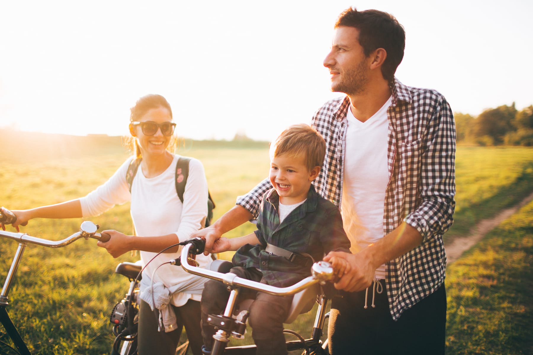 A family of three, a man, a woman, and a young child, riding bicycles together through a scenic, sun-drenched field.