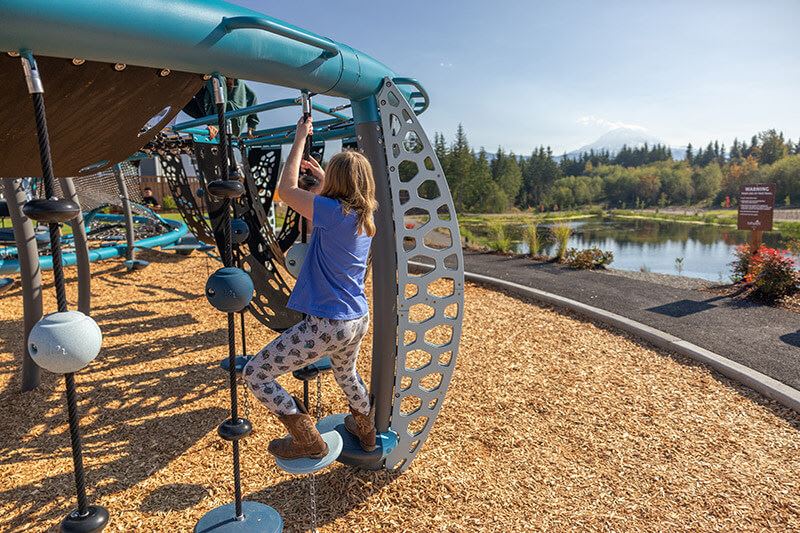 A person is climbing on a colorful, patterned play structure near a scenic lake surrounded by pine trees.