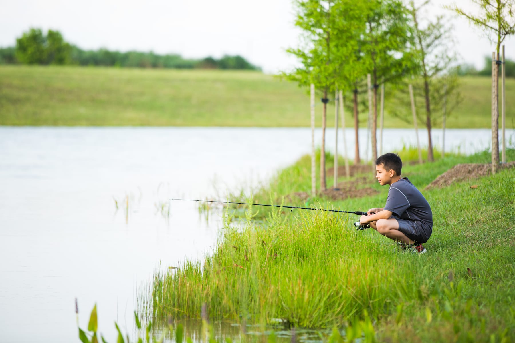 A young person is fishing by a serene lake surrounded by lush greenery and trees.